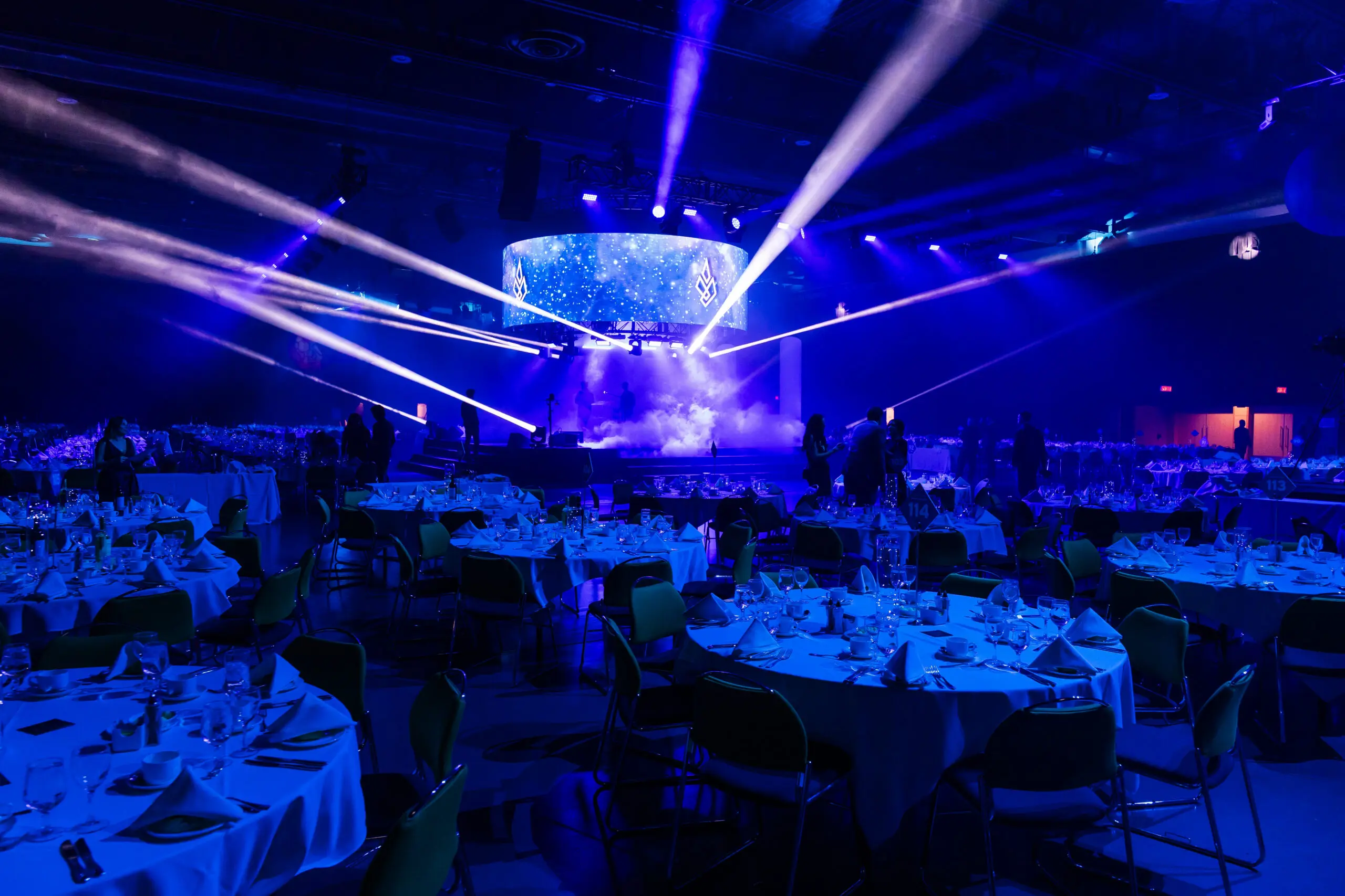 Une grande salle de banquet avec des tables rondes préparées pour le dîner, éclairée par des lumières de scène bleues et violettes. Une scène avec un écran circulaire affiche des graphiques tandis que les gens se tiennent debout et se mélangent autour de la salle.