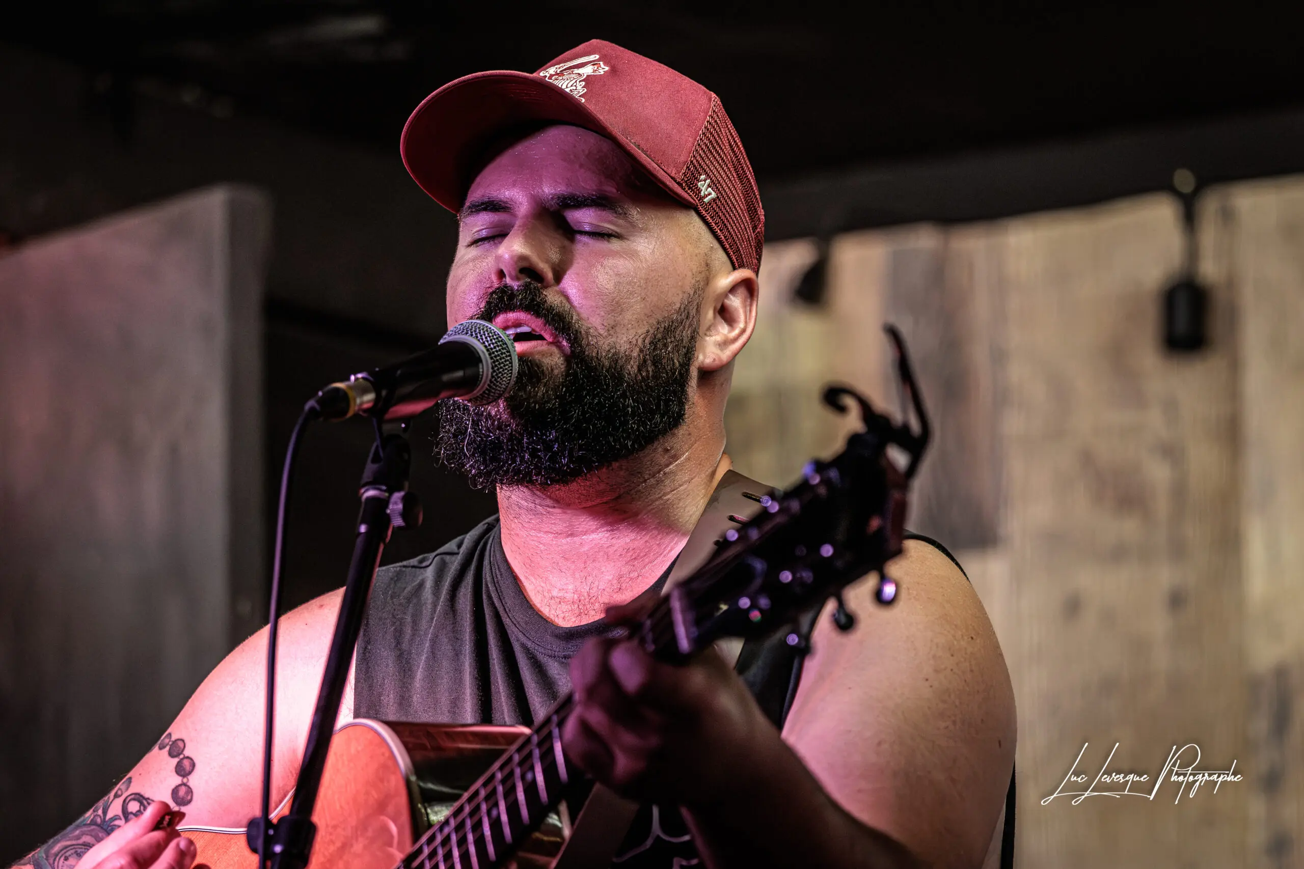 Un homme barbu portant une casquette rouge et une chemise sans manches chante passionnément dans un micro tout en jouant d'une guitare acoustique sur scène, les yeux fermés en signe de concentration.