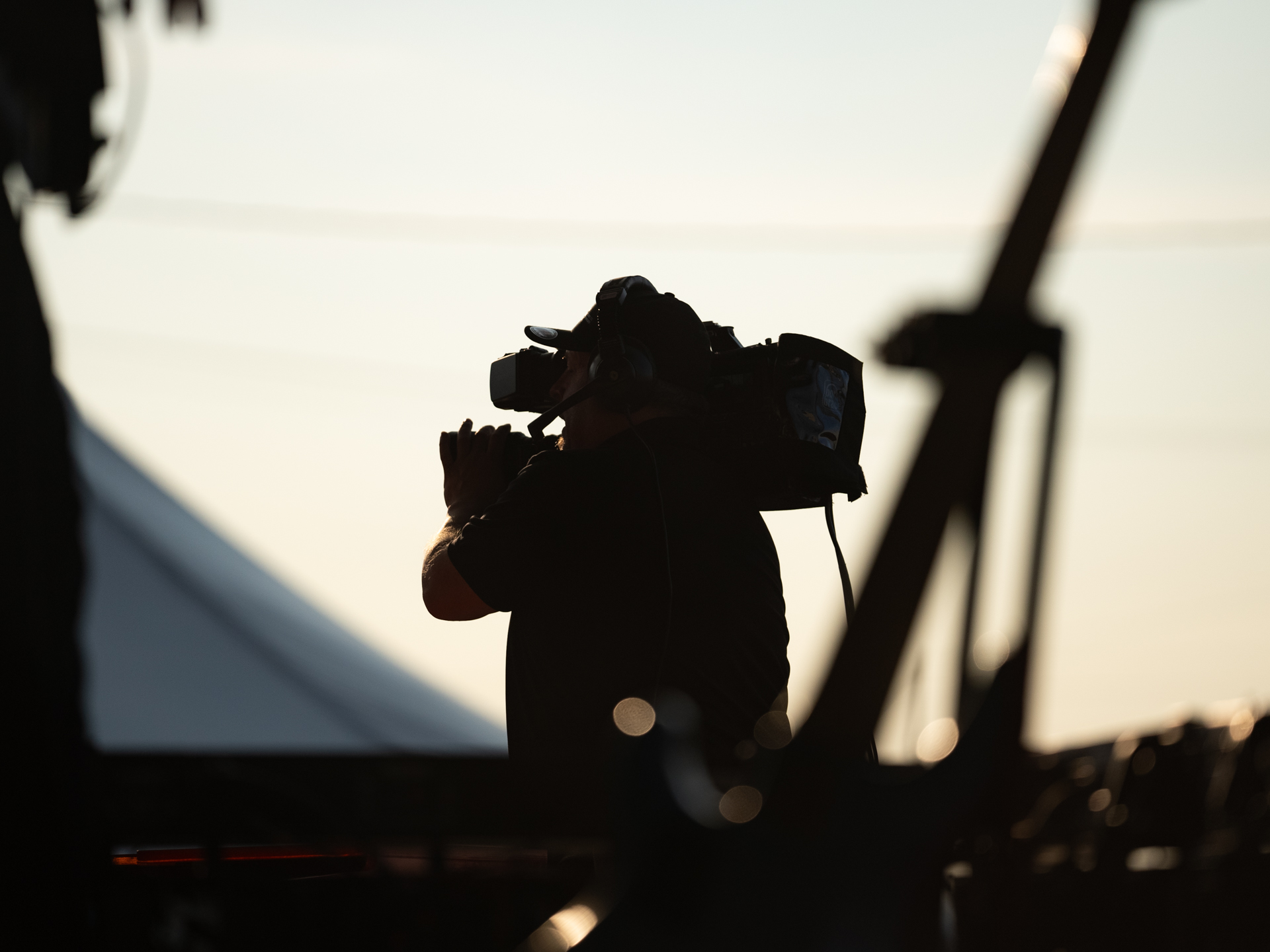 Un caméraman silhouetté enregistre un événement en plein air au coucher du soleil, avec un éclairage doux et des éléments de premier plan flous qui encadrent la scène.